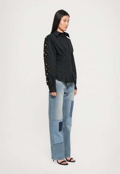 Femeie în picioare, îmbrăcată cu o bluză neagră cu puncte aurii pe mâneci, blugi deschiși la culoare cu modele patchwork și pantofi cu toc strappy negri, întorcându-se spre dreapta.