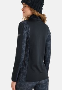 Junge Frau, die seitlich schaut, trägt einen schwarzen Rollkragenpullover mit gemusterten Ärmeln und passenden Hosen sowie eine gestrickte schwarze Mütze.