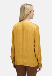 Frau mit glatten blonden Haaren, die eine senfgelbe Bluse mit langen Ärmeln und braune Hosen trägt, von hinten auf einem schlichten grauen Hintergrund zu sehen.