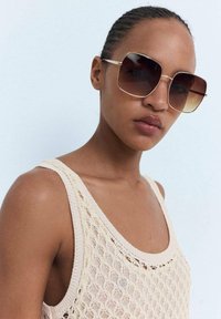 Oversized brown gradient sunglasses with a gold metal frame, featuring a square shape and large lenses. Worn with a light knitted beige top.