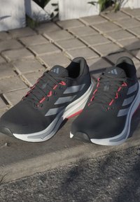 Zapatillas deportivas negras con acentos grises y rojos, que presentan una parte superior texturizada, suela acolchada y tres franjas reflectantes a cada lado.