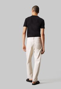 Chemise noire à manches courtes, pantalon blanc ample et sandales noires. Un fond neutre met en valeur le design et l'ajustement des vêtements.