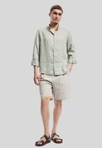 Camisa de lino verde claro de manga larga, pantalones cortos beige y sandalias marrones. Diseño simple con botones y mangas arremangadas.