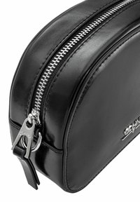 Czarna skórzana kosmetyczka o zakrzywionym kształcie, z srebrnym zamkiem błyskawicznym i wyraźnym metalowym zapięciem. Gładka tekstura, minimalna widoczna marka.