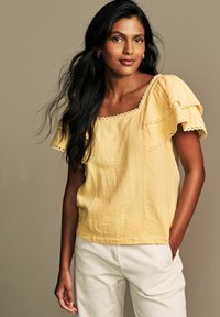 Top giallo con texture, maniche a volant e bordo in pizzo scallopato, abbinato a pantaloni bianchi. Design semplice, tessuto leggero.