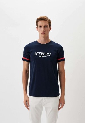 Giovane uomo che indossa una t-shirt blu navy con testo "ICEBERG BEACHWEAR" e strisce rosse sulle maniche, abbinata a pantaloni bianchi, in piedi su sfondo bianco.