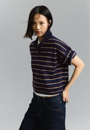 Młoda kobieta o krótkich czarnych włosach nosi granatową koszulkę polo w paski i ciemne dżinsy, pozując z jedną ręką na biodrze na szarym tle.
