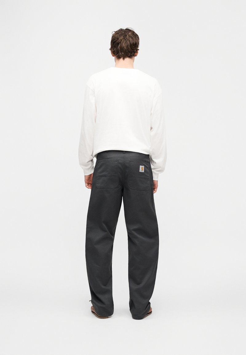 Grå arbejdsbukser med løs pasform, med en logoetiket, to baglommer og et robust stof. Kombineret med en hvid langærmet skjorte.