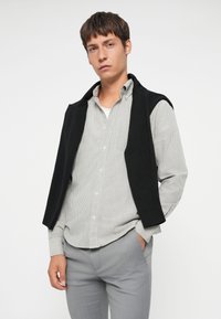 Chaleco de punto negro sobre una camisa de botones a rayas, combinado con pantalones grises claros. El conjunto presenta un diseño clásico y casual.