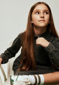 Niña con un vestido camisa de mezclilla oscuro, sentada en una silla. Cabello largo y castaño, zapatillas blancas con detalles en verde y marrón, y calcetas a rayas.