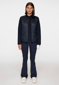 Marinblå quiltad väst med hög krage och dragkedja, tillsammans med en mörk marinblå topp och byxor. Modellen bär vita sneakers och en handske på vänster hand.