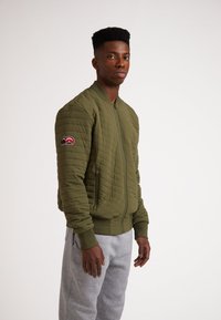 Giacca bomber imbottita verde oliva con polsini e orlo a costine, caratterizzata da una patch con logo sulla manica sinistra. Indossata sopra dei pantaloni della tuta grigi.