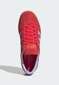 Červené semišové tenisky Adidas s bielymi pruhmi, červenými šnúrkami, gumenou podrážkou a zlatým logom Adidas na jazyku, zobrazené z horného pohľadu.