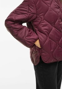 Persoană îmbrăcată într-o jachetă matlasată de culoare purpurie închisă, cu o mână în buzunarul lateralei, asortată cu pantaloni negri, pe un fundal alb.