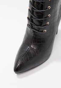 Botte noire à talon haut avec motif crocodile, bout pointu, et design à lacets avec des accents en métal rose doré. Texture lisse et brillante.