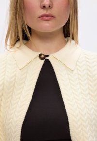 Kremowy sweter z dzianiny z teksturowanym wzorem w warkocze i płaskim kołnierzem. Zawiera okrągły brązowy guzik przy szyi.