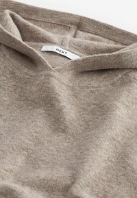 Miękki beżowy sweter z dzianiny z podwiniętym kołnierzem i widoczną metką marki "NEXT" wewnątrz dekoltu.