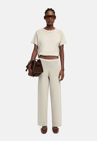 Beżowy sweter o kroju crop top i szerokie spodnie w połączeniu z brązową torebką ze zamszu. Modelka nosi duże brązowe okulary przeciwsłoneczne. Gładka tekstura na całej powierzchni.