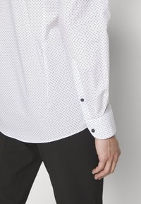 Cămașă albă cu buline navy, având o manșetă butonată și un tiv curbat. Țesătura pare netedă și ușoară, purtată cu pantaloni negri.