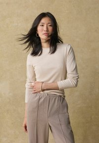 Top beige a maniche lunghe in morbido tessuto abbinato a pantaloni elasticizzati marrone chiaro con un leggero lucido e un dettaglio con bottoni.