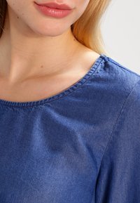 Blå denimtop med rund halsringning, slät yta och avslappnad design, med synlig sömnad längs kanterna.