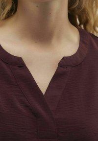 Top bordeaux a scollo a V realizzato in un tessuto leggero e testurizzato con una lucentezza sottile. Il collo presenta un bordo morbido e arrotondato.