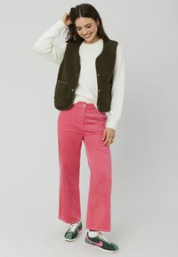Gilet en velours brun texturé sur un pull blanc ; pantalon en velours côtelé rose vif ; baskets vertes et roses ; tenue décontractée avec une coupe confortable.