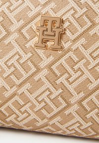 Geantă de umăr bej cu un design geometric tonal, suprafață texturată și un accent proeminent cu logo de culoare aurie în centru.