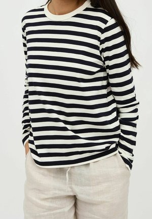 Pitkähihainen raidallinen paita, jossa on laivastonsinisiä ja beigeitä vaakaraitoja, pyöreä kaula-aukko ja rento istuvuus. Yhdistetty vaaleisiin pellavahousuihin.