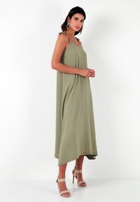 Lange olijfgroene jurk van textuurstof, met een ontspannen pasvorm, verstelbare strikbandjes en een hoge-lage zoom. Gestoeld op naakte sandalen.