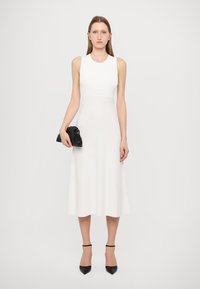 Abito midi bianco senza maniche con un corpetto aderente, che si allarga all'orlo. Abbinato a una clutch nera e a tacchi a punta neri.