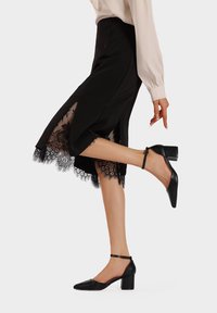 Gonna nera a linea A con bordo in pizzo, abbinata a décolleté neri con tacco blocco e cinturino alla caviglia. Tessuto fluido con una leggera lucentezza, design elegante.