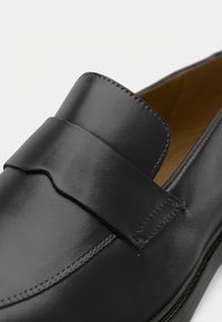 Sorte skinnloafere med en sydd rem over toppen, avrundet tå og glatt overflate. Innerringen er i beige.