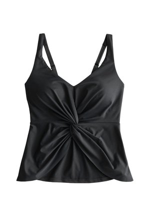 Junge Frau trägt ein schwarzes Twist-Front Tanktop und Bikinihöschen, steht draußen, lächelt und berührt mit einer Hand ihre Schulter.