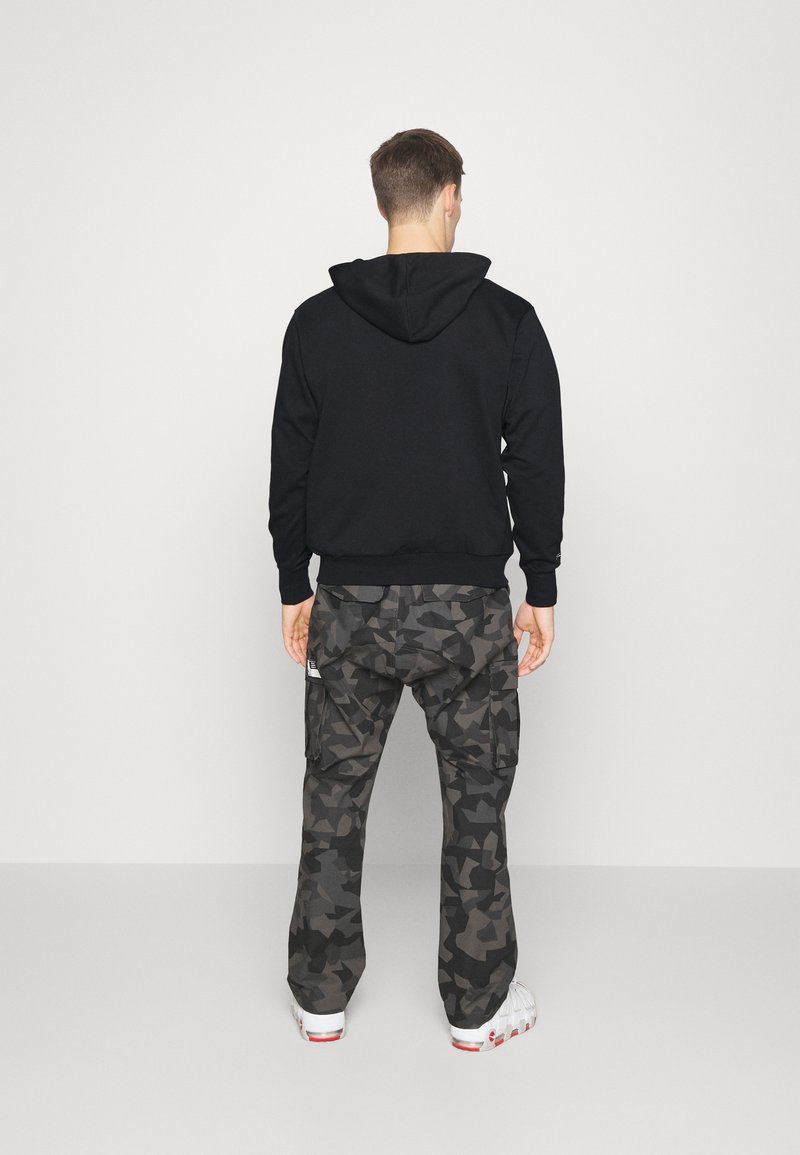 Felpa nera con cappuccio e fit flessibile, abbinata a pantaloni cargo in stile mimetico scuro con tasche laterali. Le scarpe sono bianche con dettagli rossi.