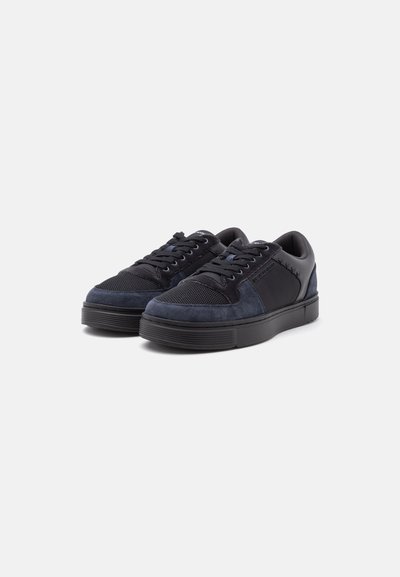 Paire de baskets basses noires et bleu foncé avec lacets, tissu en mesh, panneaux en daim et semelles épaisses en caoutchouc sur un fond clair uni.