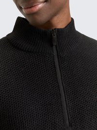 Nahaufnahme einer Person, die einen schwarzen, strukturierten Strickpullover mit einem gerippten Stehkragen und einem halbhohen Reißverschluss trägt.