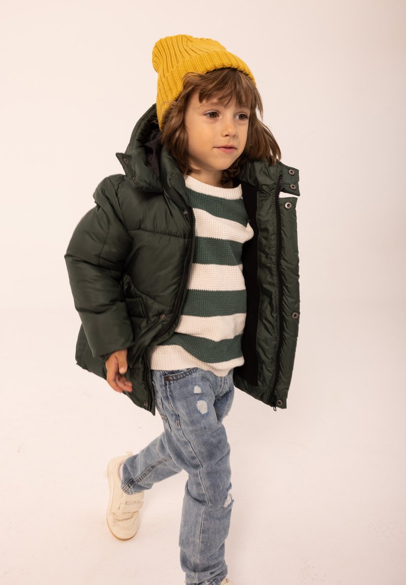 Giacca imbottita verde, maglione a strisce bianche e verdi, jeans strappati blu e berretto lavorato a maglia giallo. Sneakers bianche completano il look.