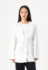 Blazer bianco texturizzato con cintura in vita, dotato di colletto a rever e due tasche frontali, indossato sopra una maglietta grafica e pantaloni neri.