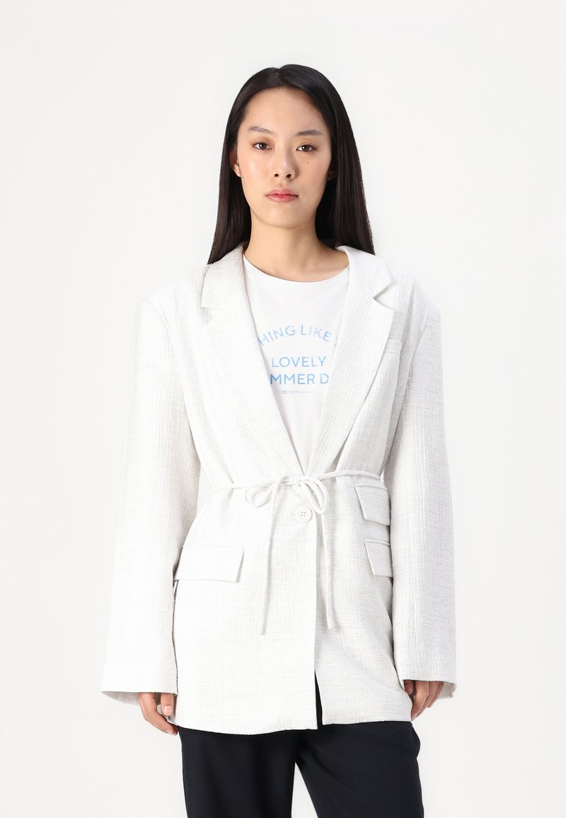 Blazer bianco texturizzato con cintura in vita, dotato di colletto a rever e due tasche frontali, indossato sopra una maglietta grafica e pantaloni neri.