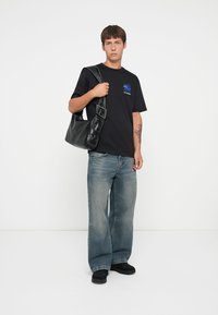 Zwarte t-shirt met een blauwe grafische print, losse blauwe jeans en een zwarte schoudertas met een glanzende textuur en gespdetails. Zwarte schoenen.