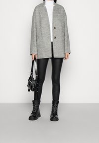 Frau trägt einen grauen Mantel mit Knöpfen, weißen Rollkragenpullover, schwarze Lederhose, schwarze Schnürstiefel und hält eine schwarze Lederschultertasche.