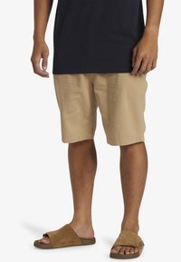 Bermudas beige de algodón con corte recto, que cuentan con bolsillos laterales. Combinadas con sandalias de ante marrones y una camiseta navy.