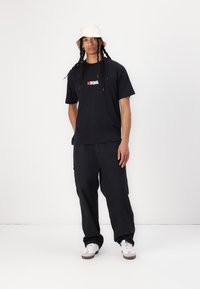 Black cotton t-shirt with logo on chest, paired with loose-fit black pants. Model wears a cream bucket hat and white sneakers.