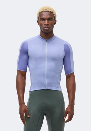 Personne athlétique aux cheveux blonds courts portant un maillot de cyclisme ajusté violet clair et un short de cyclisme rembourré vert foncé.