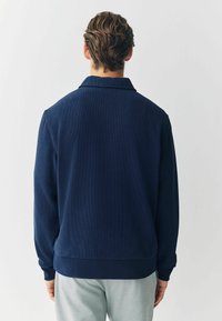 Uomo con capelli corti castani, che indossa una maglietta a maniche lunghe blu navy e pantaloni grigio chiaro, in piedi di fronte a uno sfondo bianco.
