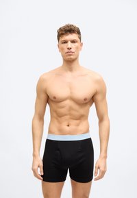 Νέος άνδρας με γυμνασμένο σώμα, φορώντας μαύρα μπόξερ Calvin Klein με ανοιχτό μπλε ζώνη στη μέση, στέκεται μπροστά σε απλό φωτεινό φόντο.