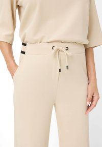 Pantaloni beige con vita elasticizzata, caratterizzati da dettagli neri e tasche laterali. Realizzati in materiale morbido e liscio.