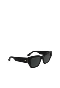 Lunettes de soleil noires avec des montures angulaires, finition mate et branches noires texturées. Présente un logo Lacoste doré et des verres sombres.