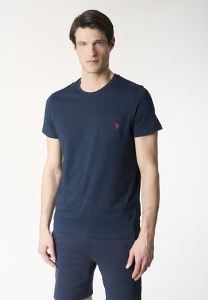 Uomo che indossa una maglietta blu navy a maniche corte con tasca e logo rosso, abbinata a pantaloncini blu navy, in piedi contro uno sfondo semplice.
